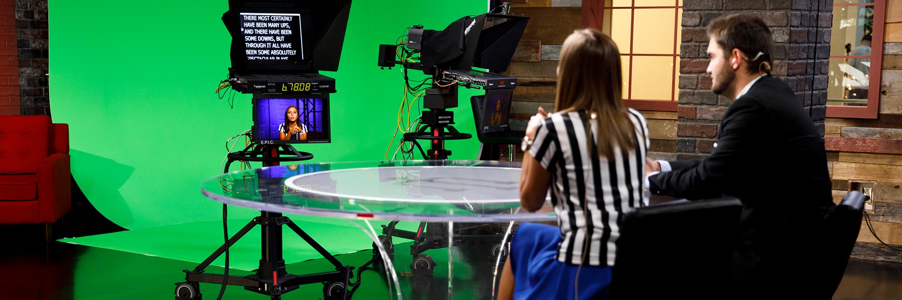 Two student reporters record a broadcast while they read from a teleprompter in the studio.