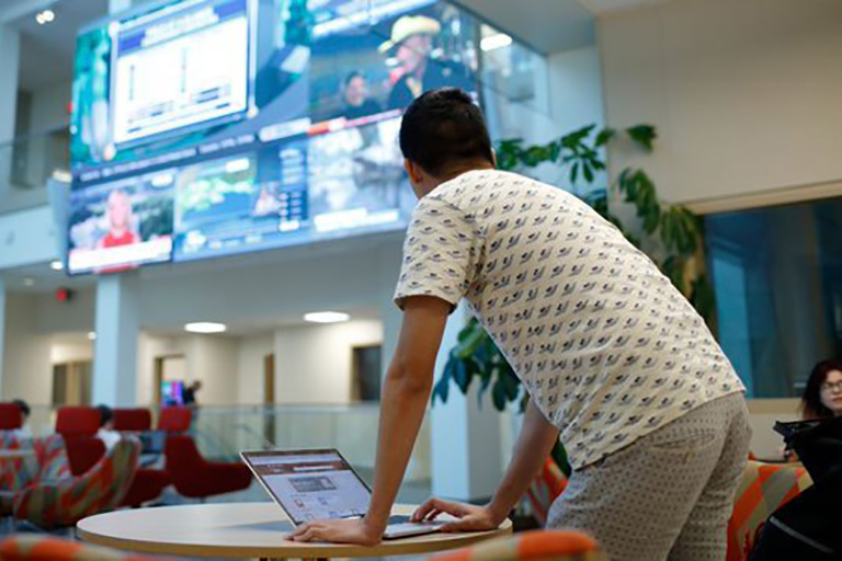 A student works on his laptop and looks to the Franklin Hall Commons big screen.
