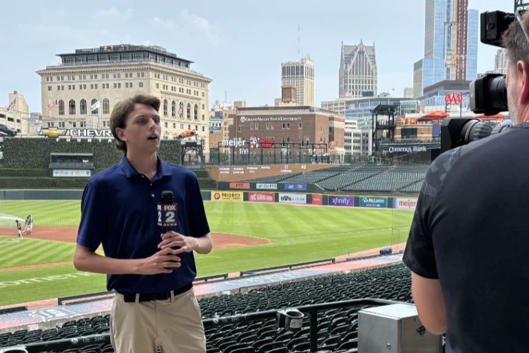 Person holding a microphone and being filmed with a baseball field and large city skyscrapers in the background.
