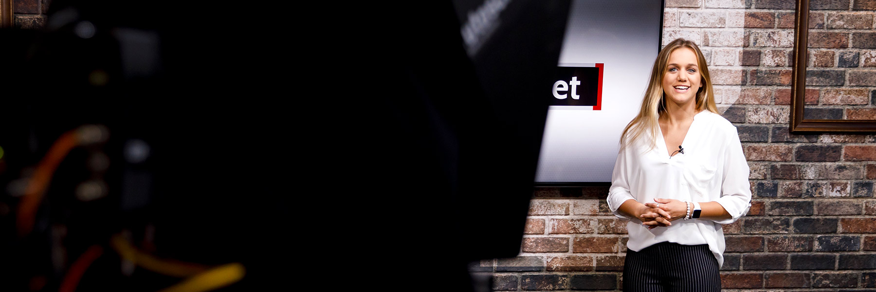 A student reports in front of a TV camera in a studio with a brick wall behind her.