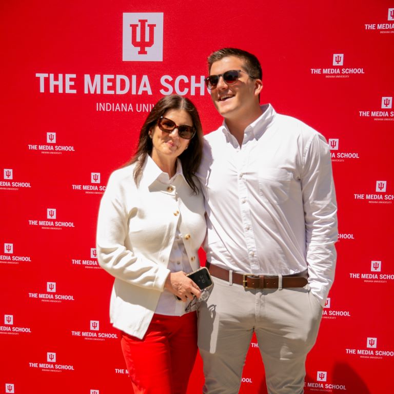 Two people stand in front of a photo backdrop with the IU logo displayed several times.